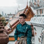Man in traditional Thai attire at a Buddhist temple with detailed architecture.