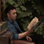 A thoughtful young man immersed in a book, surrounded by lush green plants indoors.